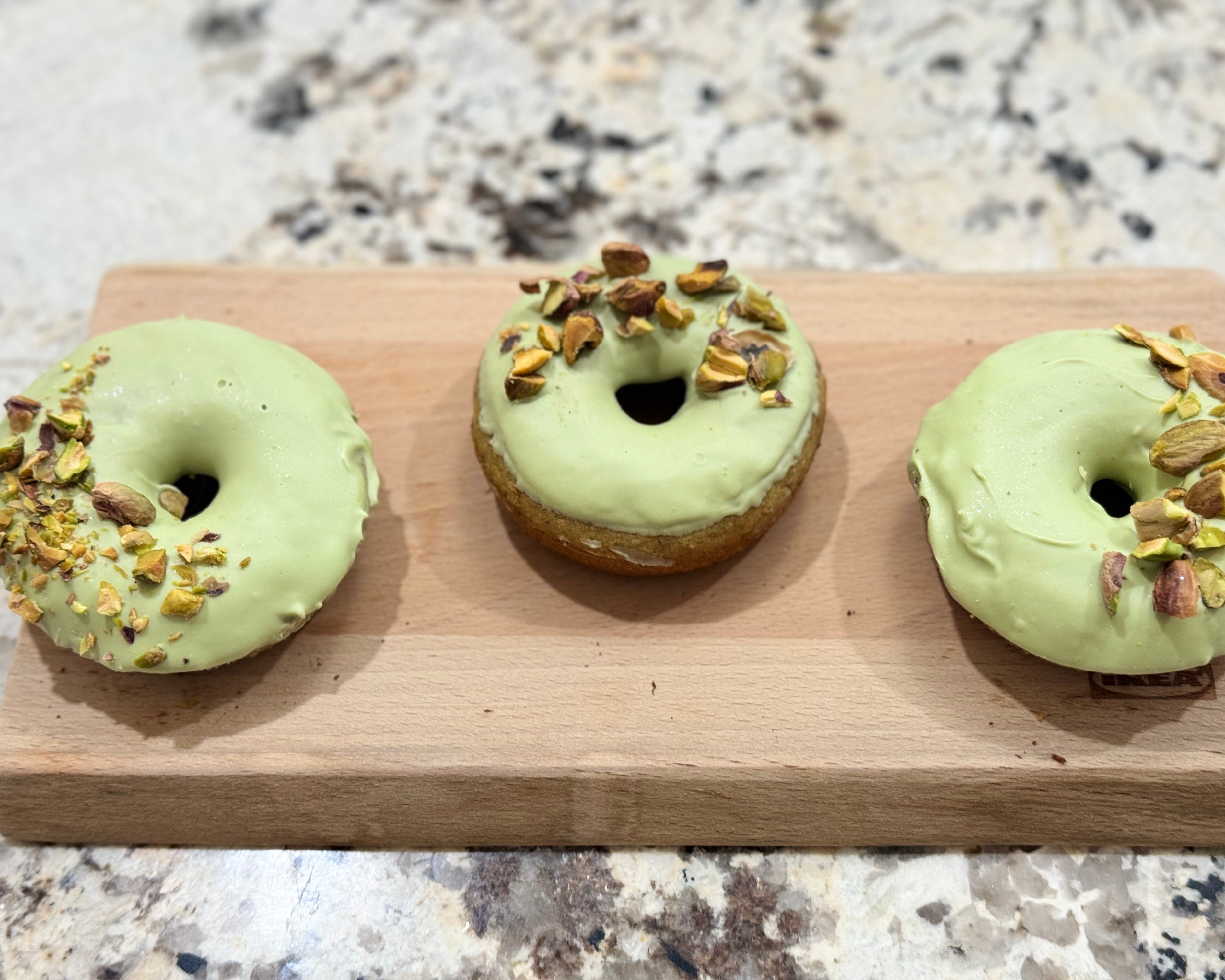 Three green doughnuts with chopped nuts on a wooden board on a marble countertop.