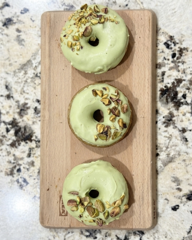 Three green doughnuts with pistachio nuts on a wooden board on a granite countertop.